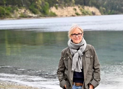 Maggie Bentley, co-founder of beanstory, wearing a grey scarf, blue sweater, and a green jacket, standing on a NW beach with the water behind her.