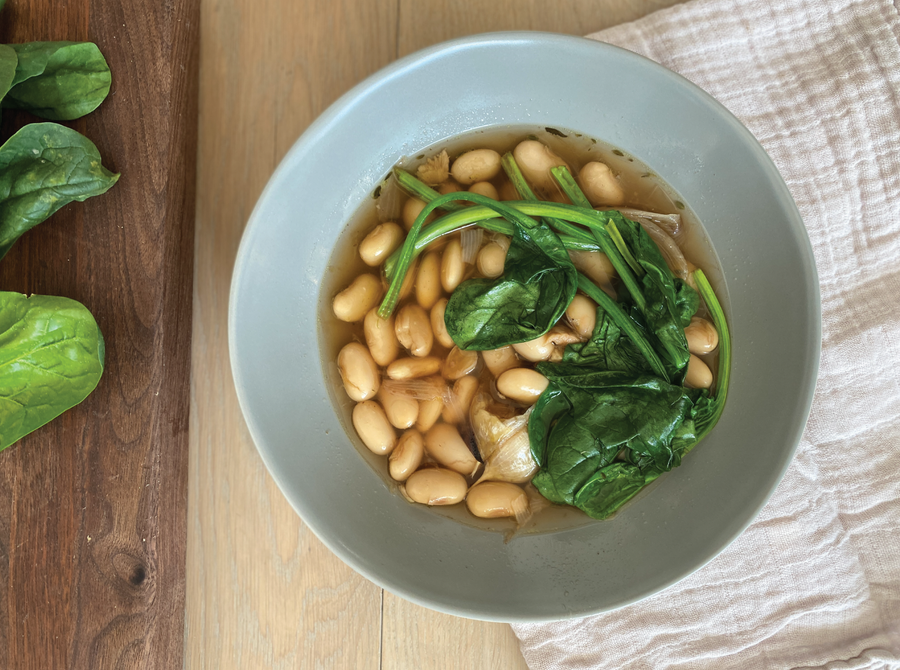 A bowl of brothy beans with wilted spinach on top, spooned into a bowl from a big pot of brothy beans made with big white runner beans and aromatics and herbs