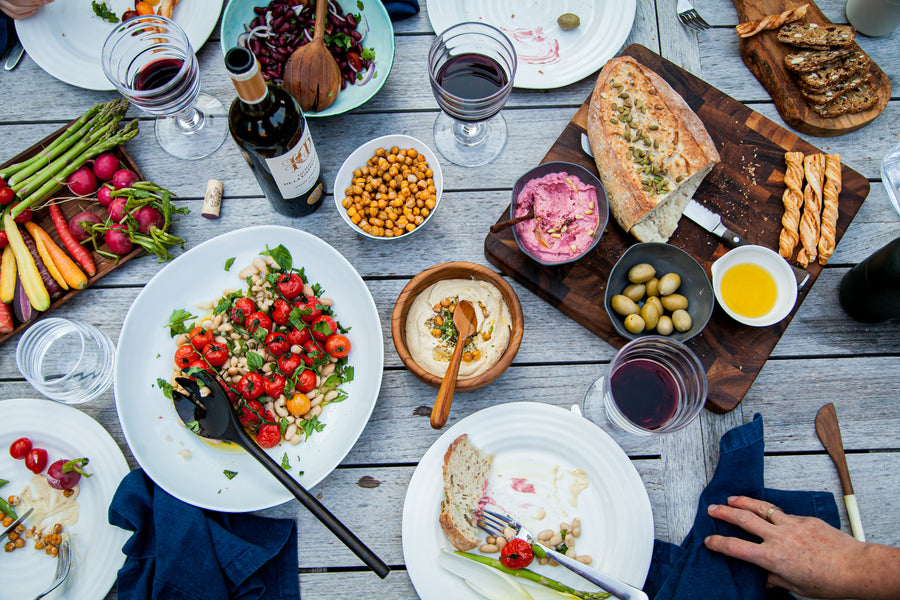 An outdoor dinner scene set on a picnic style table, filled with dishes of salads, hummus, olives, fresh bread and a delicious looking spread highlighting beanstory organic beans 