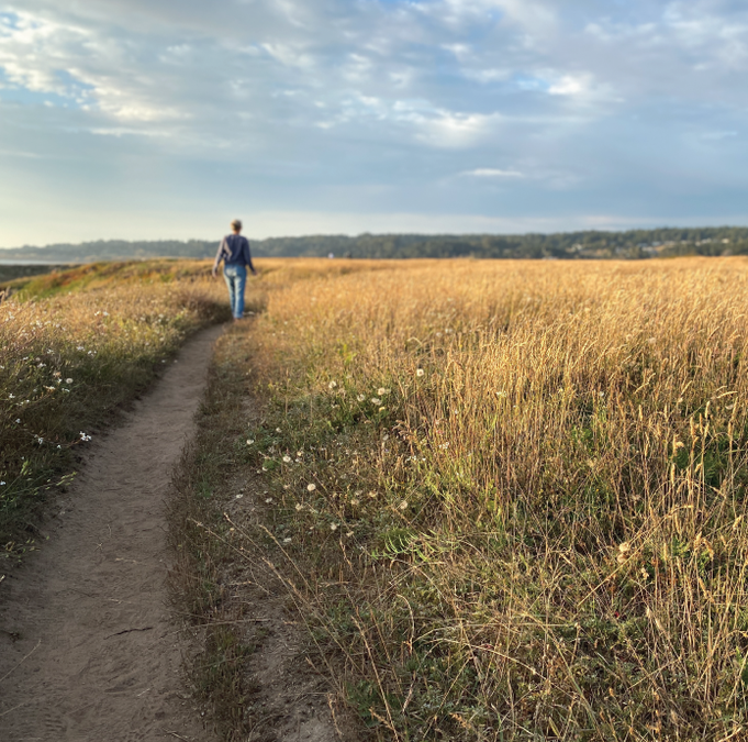 an image of co-founder, Maggie Bentley, walking along a curved sandy path bordered by tall sea grass while exploring Northern California on a visit to see farm partners on a beautiful, sunny, summer day