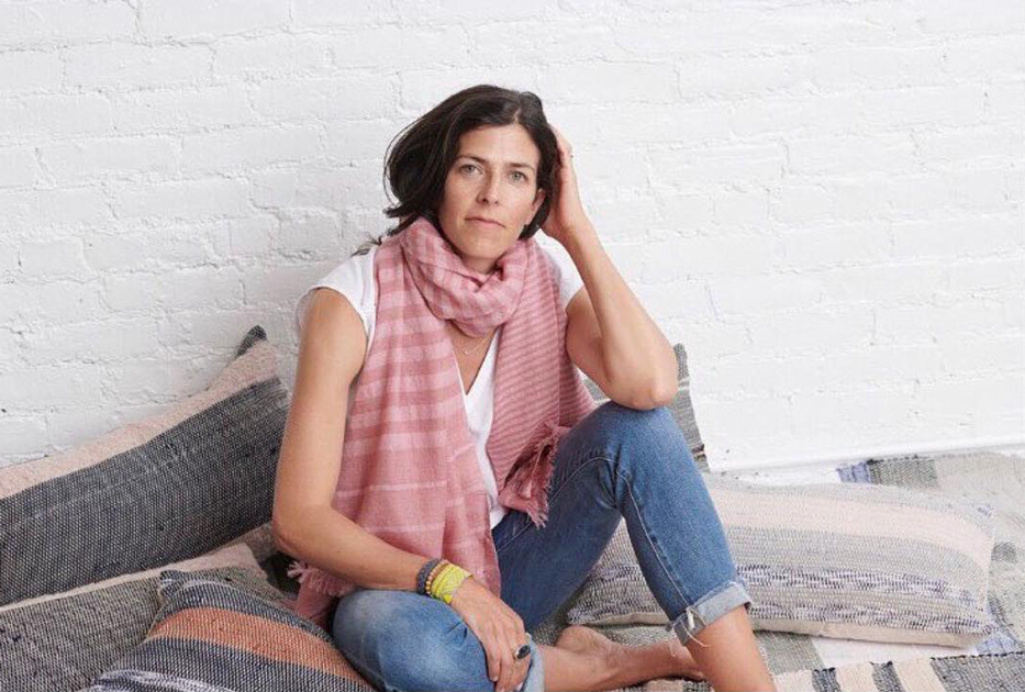 Katherine Yaphe, co-founder of beanstory, sitting on pillows that are on the floor, wearing jeans, a white shirt, and a pink striped scarf, loosely wrapped around her neck.